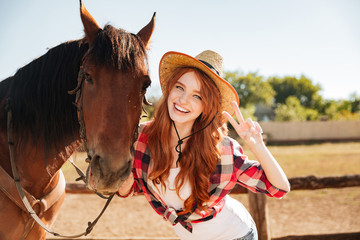Obraz premium Happy woman cowgirl standing with horse and showing peace sign