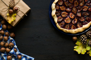 Autumn background with present box, oak leaves, acorns, pines and fruit galette pies on black wooden background. Top view.