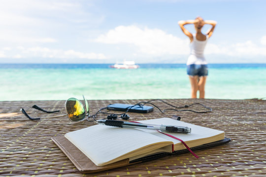 Relaxing girl, opened empty notepad is on the table with sunglasses, phone and headphones at the tropical sea background