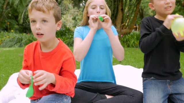 Three prepubescent children try to blow up balloons outside in yard