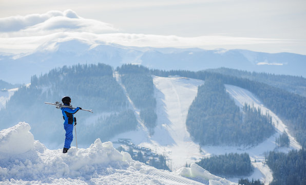 Skier standing on top of the mountain and enjoying the view on beautiful winter mountains on a sunny day. Woman is wearing blue ski suit, holding skis on her shoulder. Carpathian Mountains, Bukovel