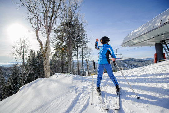 Full length portrait of female skier on the top of ski slope with ski lift and mountains on the background. Winter sports concept. Woman is looking at beautiful mountain landscape on a sunny day