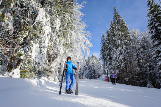 Smiling happy young female skier in winter forest. Girl is wearing blue ski suit helmet and orange glasses, holding skis. Winter sports concept. Wide angle. Carpathian Mountains, Bukovel
