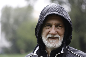 Portrait of an older white bearded man wearing a black hoodie on a rainy day with a blurry background