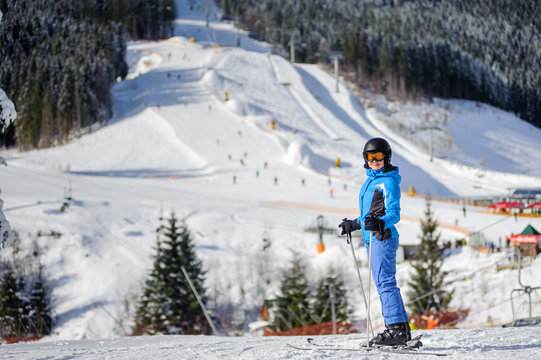 Full length portrait of happy female skier against ski slopes and ski-lift on background. Woman is wearing helmet skiing glasses gloves and blue ski suit. Winter sports concept. Carpathian Mountains