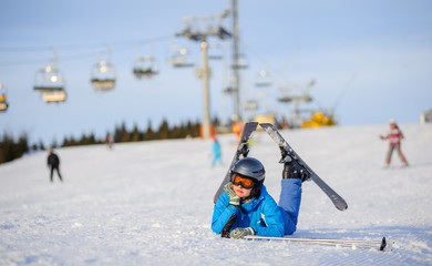 Young unhappy skier girl in blue ski suit orange goggles and helmet lying on the snow on a sunny day against ski-lift at ski resort. Winter vacation.