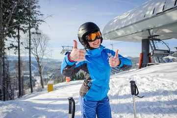 Fototapeta na wymiar Portrait of young beautiful female skier at ski resort smiling and showing thumbs up. Winter sports concept. Woman is wearing blue jacket and blue pants, helmet and orange goggles. Bukovel, Ukraine