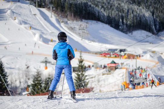 Rear view of skier standing on ski slope getting ready to ski against ski resort on background. Girl is wearing helmet and blue ski suit. Winter sports concept. Carpathian Mountains, Bukovel
