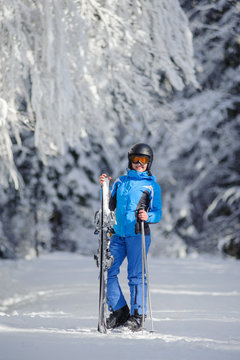 Full length portrait of young happy female skier standing enjoying sunny day against beautiful snow covered trees on the background. Woman is holding her skis in one hand and ski poles in another.