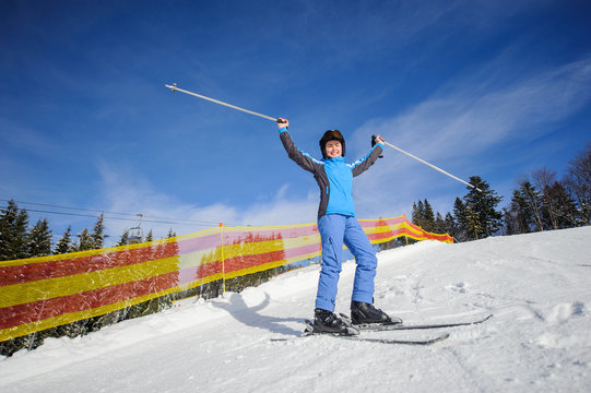 Happy young female skier on a sunny day at ski resort. Woman raised her arms as sign of success. Winter sports concept.