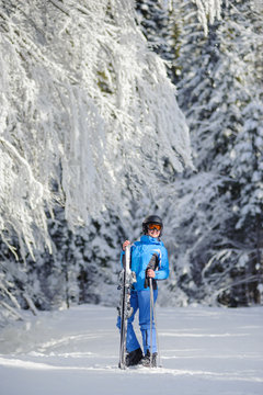 Happy female skier standing and enjoying sunny day against beautiful snow covered trees on the background. Girl is holding her skis in one hand and ski poles in another. Carpathian Mountains, Bukovel