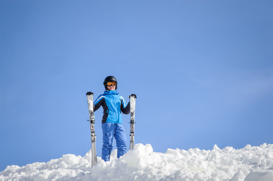 Portrait of young female skier wearing blue ski suit, helmet and goggles on sunny day. Woman is holding her skis. Winter sports concept.
