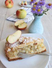 Apfelkuchen auf einem weißen Teller mit frischen Äpfeln, Blumen und einer Tasse Kaffee im Hintergrund auf einem grauen Holztisch