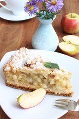 Apfelkuchen auf einem weißen Teller mit frischen Äpfeln, Blumen und Kaffee im Hintergrund auf einem braunen Holztisch