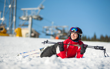 Fototapeta na wymiar Smiling girl lying with skis on snowy at mountain top, holding ski-sticks and looking away in sunny day at a winter resort with ski lifts and blue sky in background. Close-up