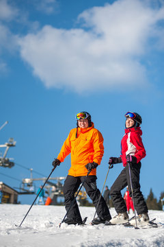 Couple holding skis smiling on mountain top together at a winter resort with ski lifts and blue sky in background. Man is wearing orange jacket, female in red jacket, both is wearing helmets