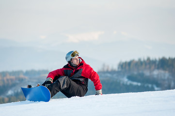 Fototapeta na wymiar Snowboarder wearing helmet, red jacket, gloves and pants sitting on snowy slope on top of a mountain looking away, with an astonishing view on hills. Carpathian mountains