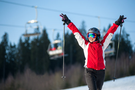 Smiling female skier wearing helmet, red jacket and ski goggles standing on snowy slope with hands raised up in sunny day with forest and blue sky in background. Close-up
