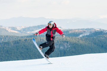 Fototapeta na wymiar Cool man snowboarder riding on his snowboard and taking his for the edge on top of a mountain against the backdrop of mountains, hills and forests in the distance.