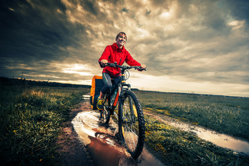 Fototapeta premium Young lady hiker with loaded bicycle riding through the puddle on a wet rural road