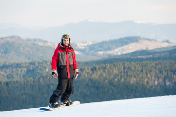 Fototapeta na wymiar Young snowboarder wearing helmet, red jacket, gloves and pants standing on top of a mountain and looking at the camera on the background of beautiful mountains and forests, winter sports concept