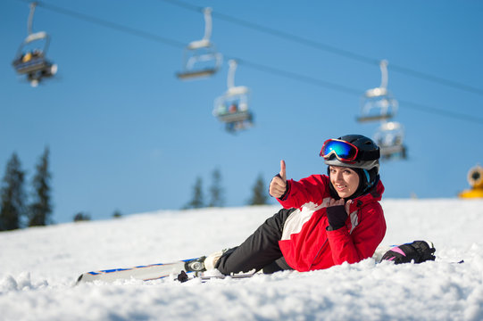 Smiling girl lying with skis on snowy at mountain top, showing thumb up gesture of good class and looking at the camera in sunny day at a winter resort with ski lifts and blue sky in background