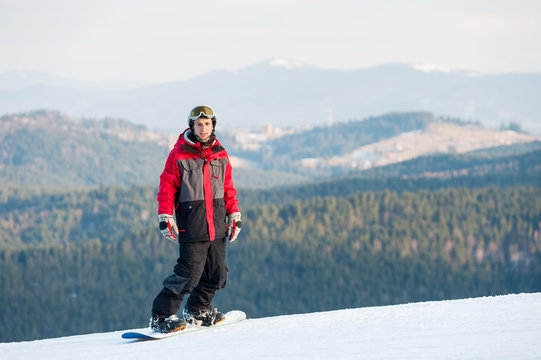 Young snowboarder wearing helmet, red jacket, gloves and pants standing on top of a mountain and looking at the camera on the background of beautiful mountains and forests, winter sports concept