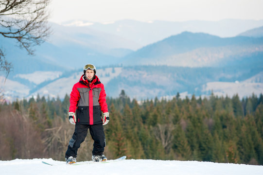 Portrait of snowboarder wearing helmet, red jacket, gloves and pants standing on top of a mountain and looking at the camera on the background of forests, hills and the sky