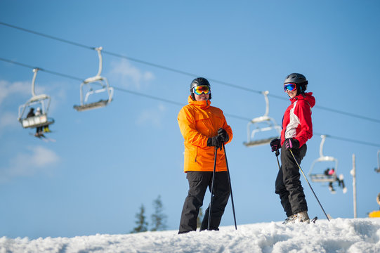 Couple male and female wearing ski goggles standing with skis on mountain top at a winter resort in sunny day with ski lifts and blue sky in background.