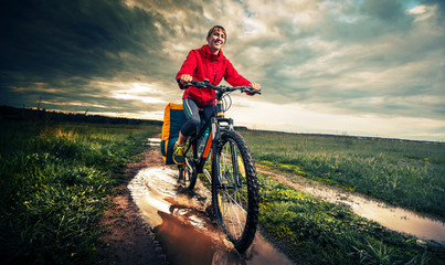 Young lady hiker with loaded bicycle riding through the puddle on a wet rural road