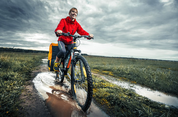 Young lady hiker with loaded bicycle riding through the puddle on a wet rural road