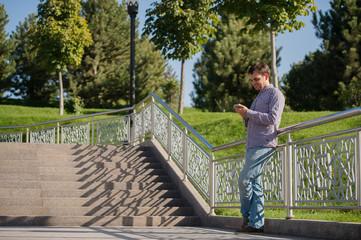 Young hispanic man, blue shirt and jeans, standing on stairs outdoors, holding mobile phone in his hand, smiling - communication concept