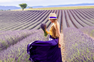 Young pretty woman in the long waving skirt and hat standing with lavender bouquet on the lavender...