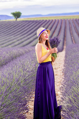 Young and pretty woman in the long violet skirt standing with lavender bouquet on the lavender field in Provence in France