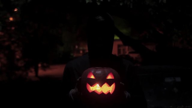 Black silhouette figure carries jack o lantern carved pumpkin through dark alley