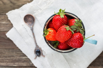 Fresh juicy strawberries in old rusty mug. Rustic wooden background with hand made napkin and vintage spoon. Top view, place for text.