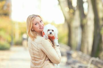 Young woman is holding her cute Maltese terrier and looking at camera over the shoulder. They are walking in a park on a beautiful autumn day.