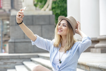 Fototapeta premium Beautiful woman tourist making selfie on the steps of the historic building. Girl blonde, wearing a white T-shirt, blue shirt and blue denim shorts, a hat on her head. Summer. Outdoors.