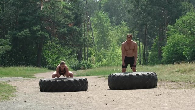 Slow motion shot of fitness couple flipping large tractor tires towards the camera on dusty road