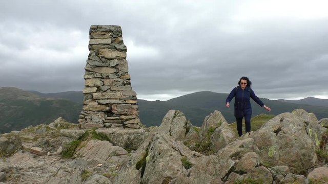 Peak of the mountain in Lake District, England, UK.Smiling woman in strong wind climbing to the summit of mountain and reaching stone cairn