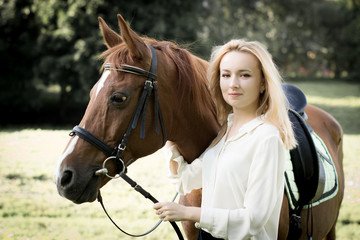 Молодая девушка со светлыми волосами стоит рядом с коричневой лошадью 