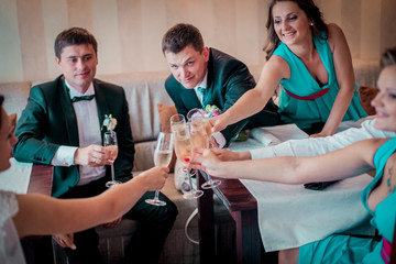 Stylish newlyweds dring champagne with their friends sitting in hotel room or cafe.
