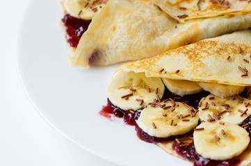 Thin pancakes with banana slices, sweet cherry jam and chocolate flavoured strands on white plate. Breakfast meal. Dessert food