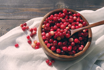 Fresh red forest cranberry in a round bowl with a wooden spoon on white linen fabric on a table surface. Autumn harvest of wild berries