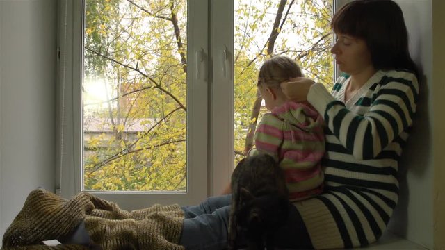 Мать заплетает дочери волосы , сидя на подоконнике в осенний пасмурный день. Маленькая девочка гладит кошку.