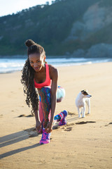 Sporty woman lacing sport footwear before running and exercising at the beach with her dog. Black beautiful runner training outdoor.