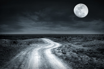 Empty rural road going through prairie at full moon night with dramatic cloudy sky