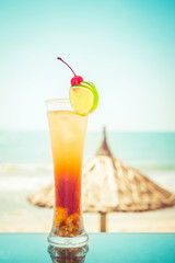 Long Island cocktail with fruits decoration at tropical ocean beach with umbrella. Vintage style, hipster colors image with copy space for party invitation text