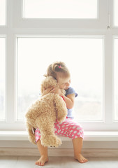 Adorable little girl hugging a teddy bear. Cute baby at home in white room is sitting near window.