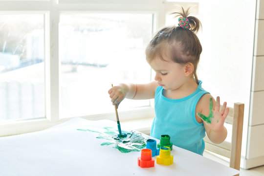 Cute little toddler child painting with paintbrush and colorful paints. Adorable baby girl drawing on white paper near window in light room